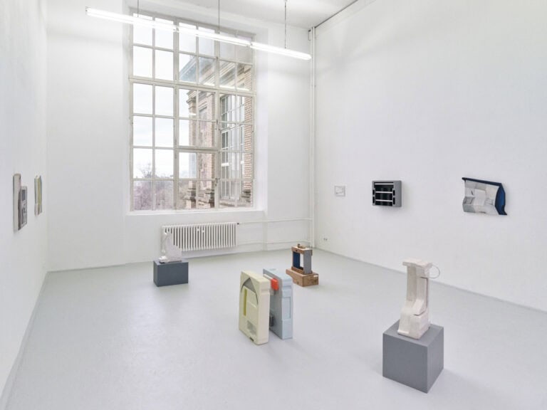 Foto: Ansicht eines Raumes mit weißen Wänden, grauem Boden und großem Fenster. An den Wänden befinden sich Objekte im Klein- und Mittelformat. Im Raum befinden sich weitere Objekte, die zum Teil auf Sockeln stehen.