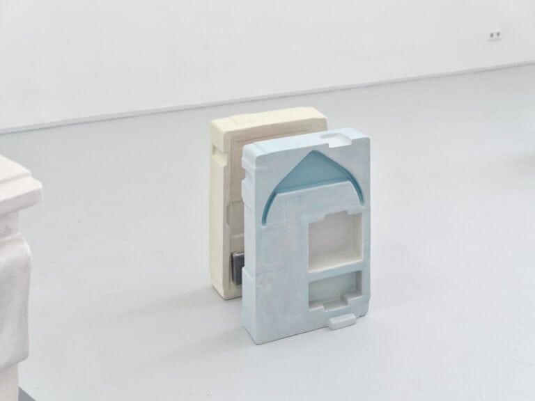 Foto: Nahaufnahme. Ein Raum mit weißen Wänden und grauem Boden. Auf dem Boden stehen zwei Objekte mit rechteckiger Grundform.