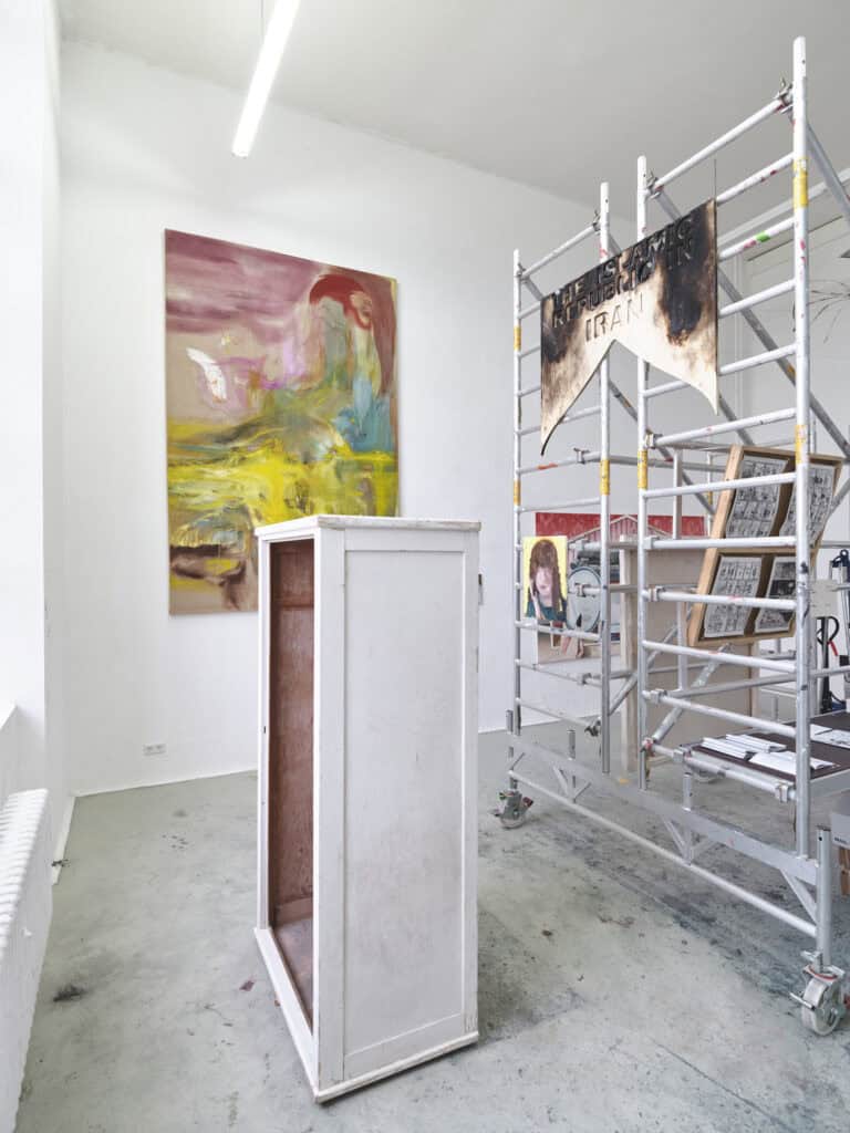 Foto: Ansicht eines Raumes mit weißen Wänden und grauem Boden. Wände und Boden sind teilweise mit Farbspuren versehen. Im Raum befindet sich ein kastenartiges Objekt und ein Baugerüst. Auf den Streben und Plattformen des Gerüsts sind Malereien und Skulpturen angebracht sind. An den Wänden befinden sich Malereien.