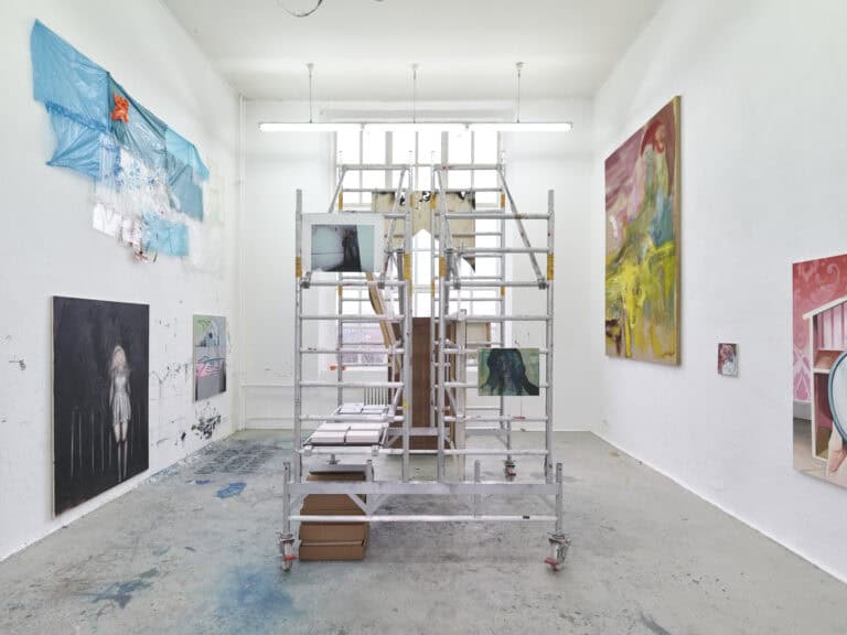 Foto: Ansicht eines Raumes mit weißen Wänden und grauem Boden. Wände und Boden sind teilweise mit Farbspuren versehen. In der Mitte des Raumes befindet sich ein Baugerüst, auf dessen Streben und Plattformen Malereien und Skulpturen angebracht sind. An den Wänden befinden sich klein- bis großformatige Malereien und Objekte.