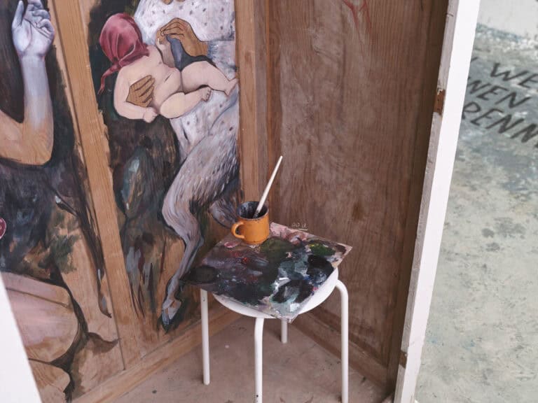 Foto: Nahaufnahme. Das Innenleben einer einseitig geöffneten Holzbox. Eine Seite der Box ist bemalt. In der Box steht ein Hocker, auf dem sich eine Tasse, eine Malpalette und ein Pinsel befinden. Im Hintergrund ist der graue, zum Teil mit Flecken versehene Raumboden zu sehen, auf den Buchstaben aufgebracht sind.