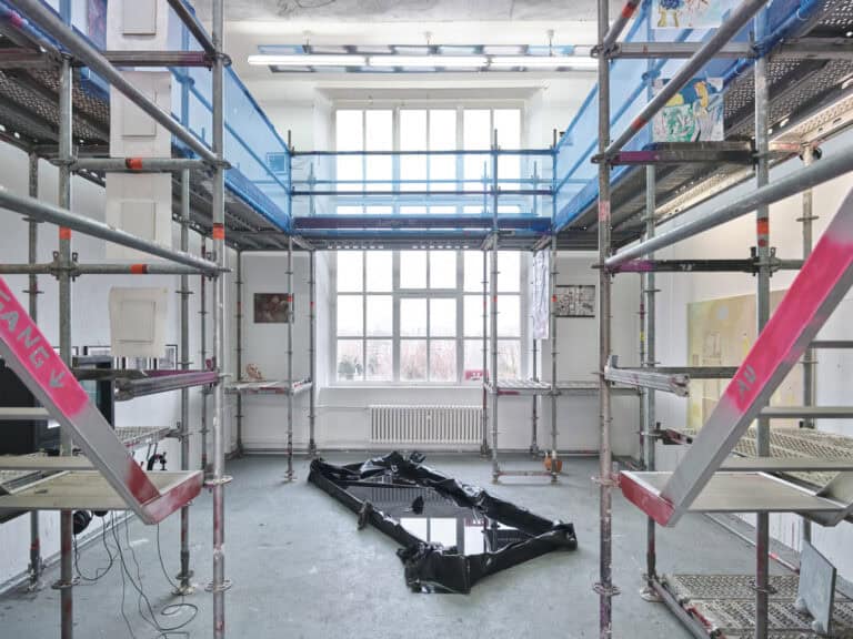 Foto: Ansicht eines Raumes mit weißen Wänden, grauem Boden und einem großen Fenster. Im Raum befindet sich ein Baugerüst auf Metall mit Treppenaufgang. Zwischen den Streben des Gerüstes und an der Wand sind Malereien und Papierarbeiten angebracht. Auf dem Gerüst stehen Skulpturen und ein Fernsehen. Der Oberste Etage des gerüstet ist mit einem Netz versehen. In der Mitte des Raumes befindet sich ein mit Wasser gefülltes Becken aus schwarzer Folie, in dem schwarze Schuhe liegen.