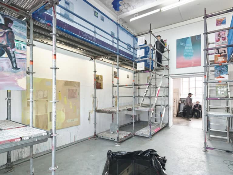 Foto: Aufnahme eines Raumes mit weißen Wänden und grauem Boden. Im Raum befindet sich ein mehrgeschossiges, begehbares Baugerüst, auf dem sich Menschen befinden. Zwischen den Streben des Gerüstes und an der Wand sind Malereien, Objekte und Papierarbeiten angebracht. An der Decke ist eine Deckenbemalung.