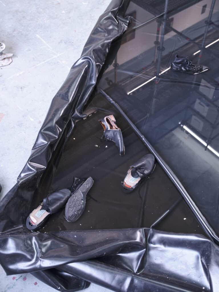 Foto: Nahaufnahme eines Beckens aus schwarzer Folie. In dem Becken befindet sich mehrere Zentimeter tiefes Wasser und mehrere schwarze Schuhe. Das Becken liegt auf einem grauen Boden.