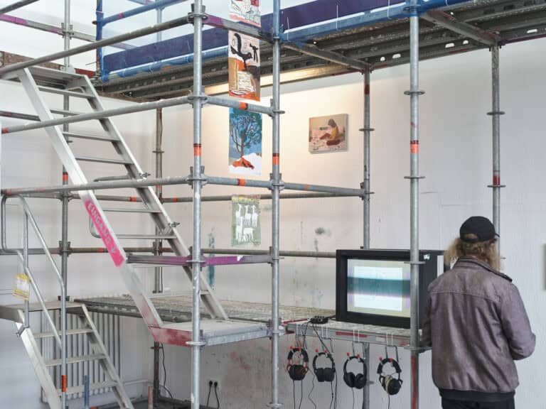Foto: Ansicht eines Raumes mit weißen Wänden. Die Wände sind zum Teil mit Farbspuren versehen. Im Raum befindet sich ein Baugerüst aus Metall mitsamt einer Treppe und mehreren, begehbahren Ebenen. Zwischen den Streben des Gerüstes und an der Wand sind Malereien und Papierarbeiten angebracht. Auf dem Gerüst steht ein Fernsehen, vor dem mehrere Kopfhörer hängen. Rechts im Bild steht ein Mensch.