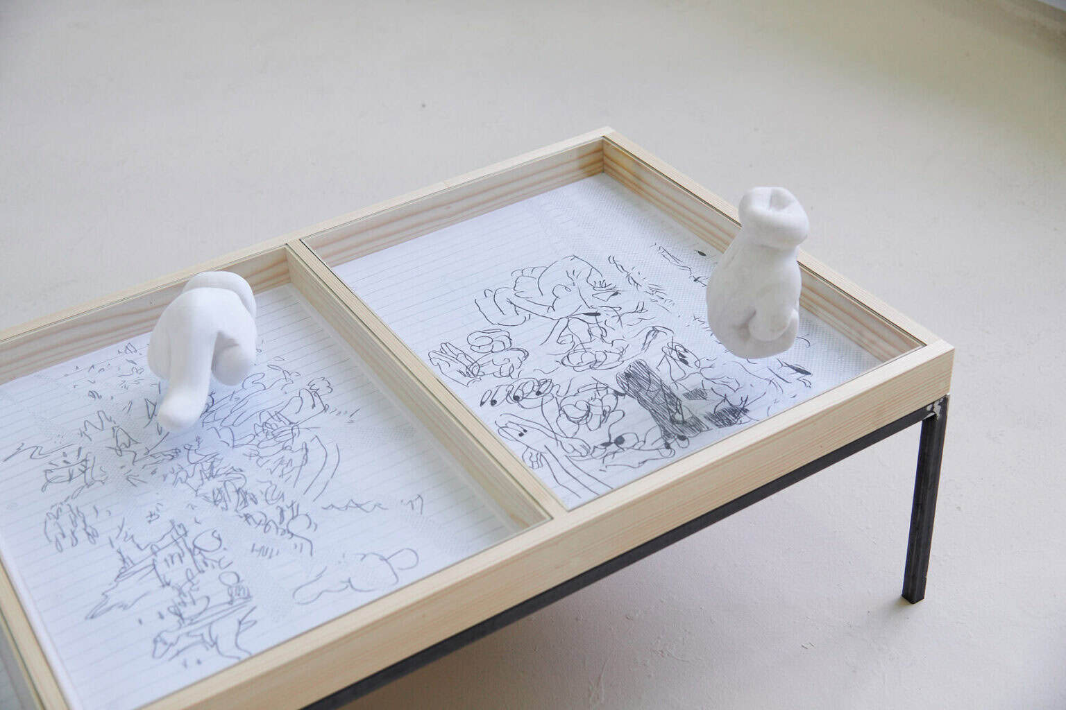 Foto: Ein Raum mitbeigem Boden. Im Raum steht eine Vitrine. In ihr liegen Papierarbeiten. Auf dem Glas der Vitrine, über den Zeichnungen, liegen zwei Objekte.