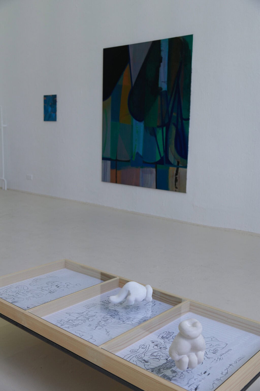 Foto: Ein Raum mit weißen Wänden und beigem Boden. Im Raum stehen zwei Vitrinen. In einer von ihnen liegen Papierarbeiten. Auf dem Glas der Vitrine, über den Zeichnungen, liegen zwei Objekte. An einer der Wände hängen zwei Leinwandarbeiten.