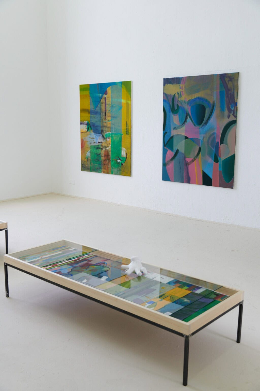 Foto: Ein Raum mit weißen Wänden und beigem Boden. Im Raum stehen zwei Vitrinen. In einer von ihnen liegen Papierarbeiten. Auf dem Glas der Vitrine, über den Arbeiten, liegt ein Objekt. An einer der Wände hängen drei großformatige Leinwandarbeiten.