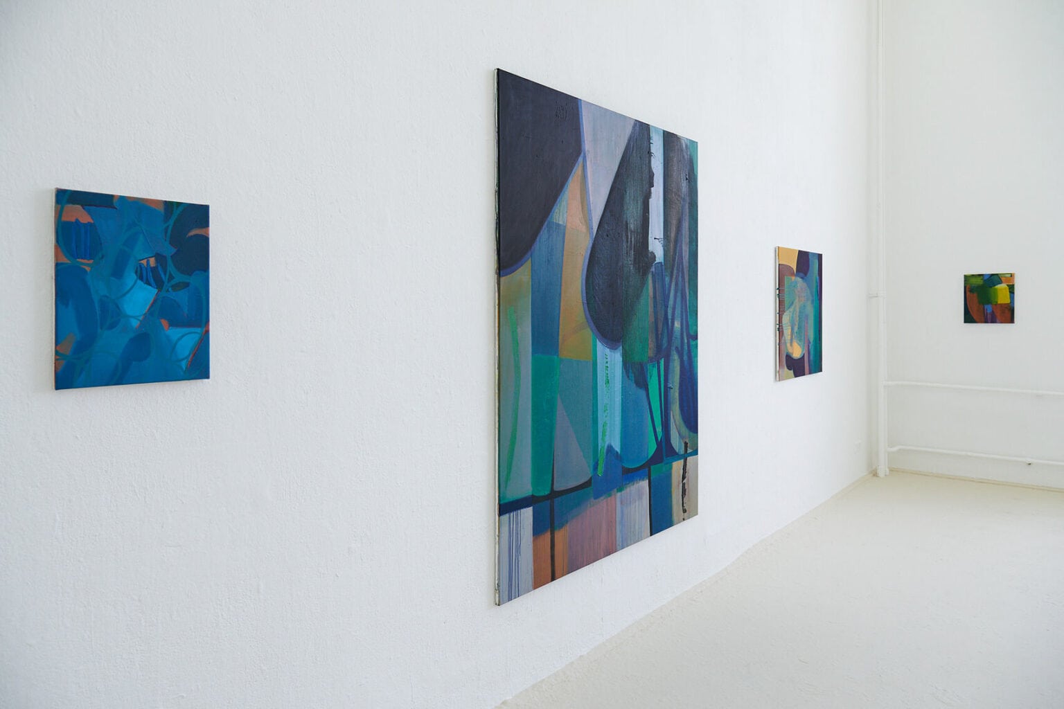 Foto: Ein Raum mit weißer Wand und beigem Boden. An der Wand hängen klein-, mittel- und großformatige Leinwände.