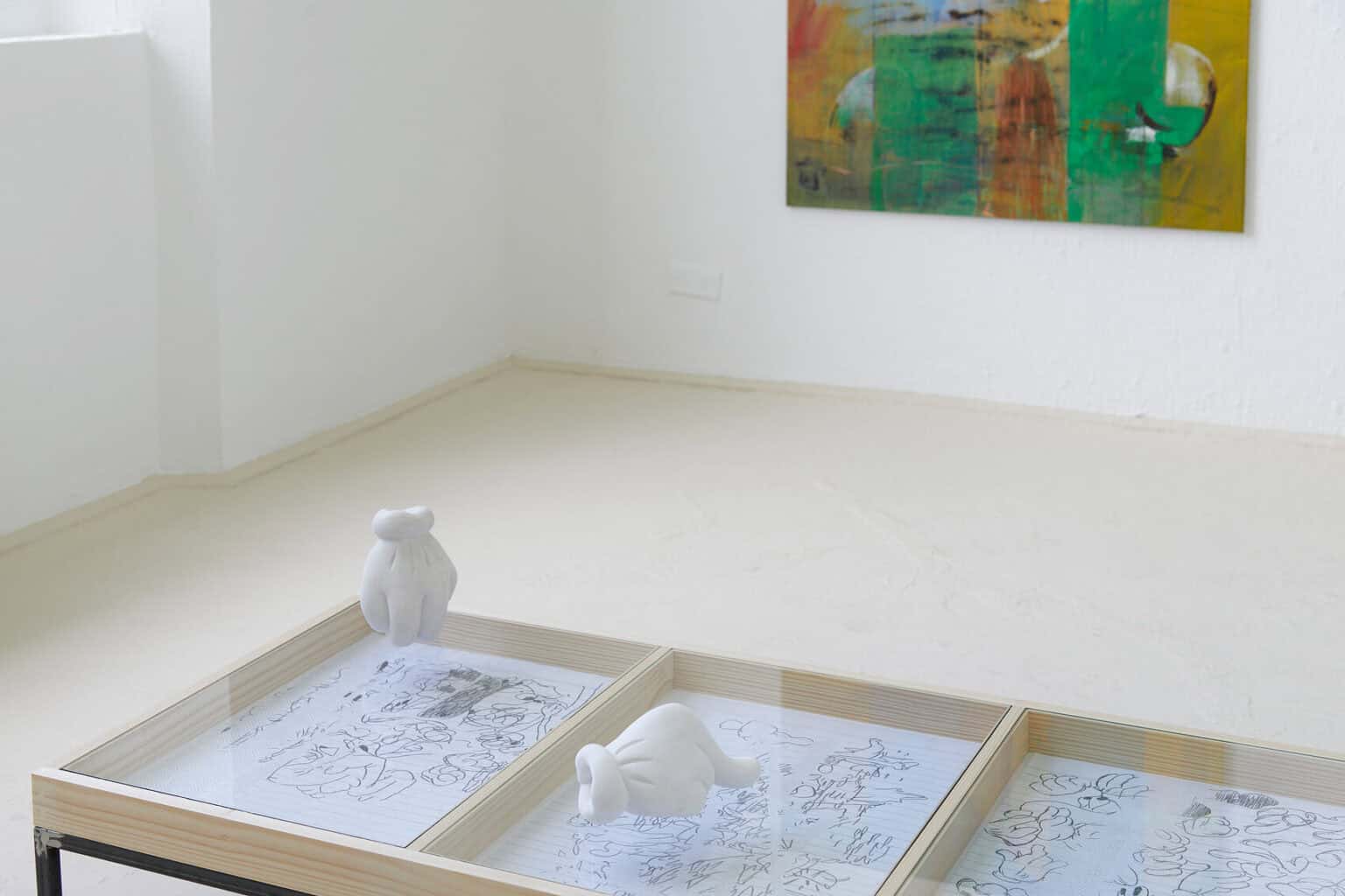 Foto: Ein Raum mit weißen Wänden und beigem Boden. im Raum steht eine Vitrine, in der Papierarbeiten liegen. Auf dem Glas der Vitrine, über den Zeichnungen, liegen zwei Objekte. An einer der Wände hängt eine Leinwand.