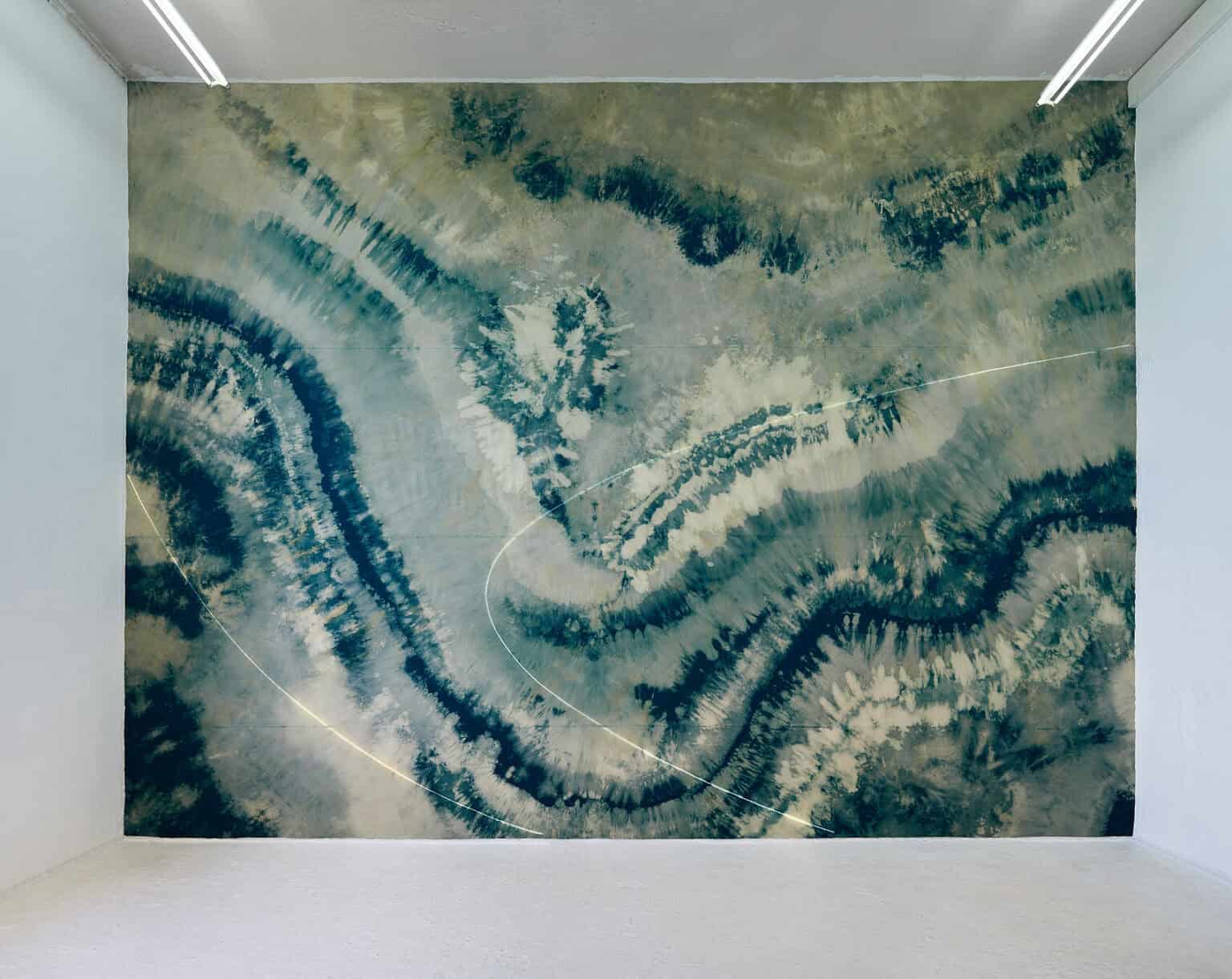 Foto: Ansicht eines Raumes mit weißen Wänden und hellgrauem Boden. Eine der Wände ist in Gänze mit einem bunten Material behangen.