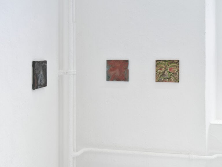 Foto: Nahaufnahme der Ecke eines Raumes. Weiße Wände mit weißen Rohren. An der Wand befinden sich drei Malereien im gleichen Format, auf gleicher Höhe.