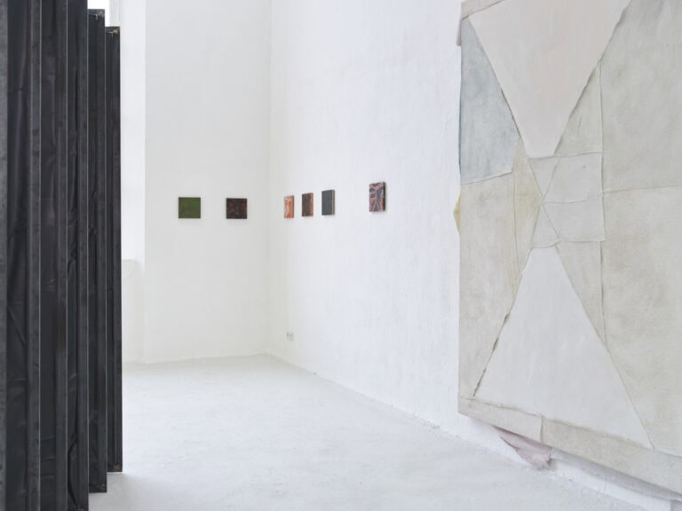 Foto: Ansicht eines Raumes mit weißen Wänden und einem grau-weißen Boden. Links im Bild zu sehen ist eine schwarzes Objekt. An den Wänden hängen klein- und großformatige Objekte.