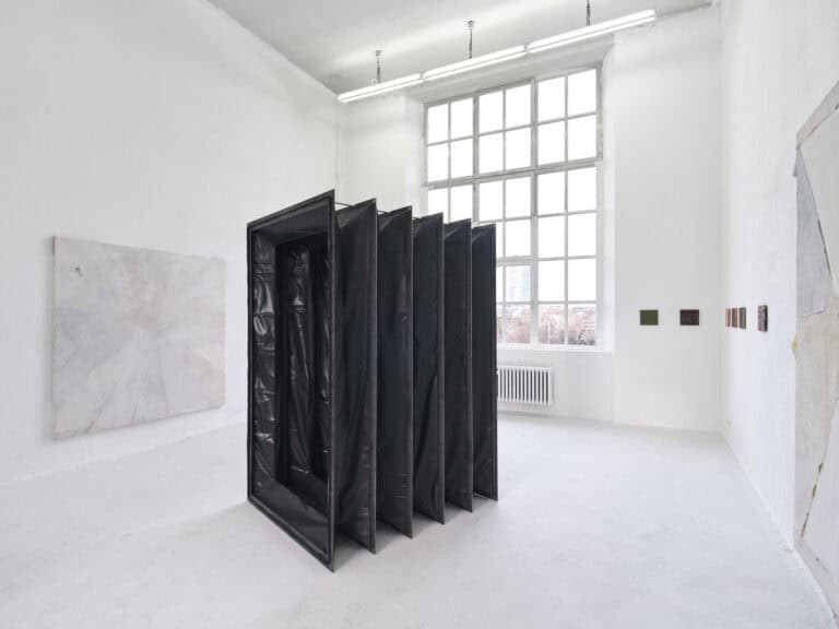 Foto: Ansicht eines Raumes mit weiß-grauem Boden. An den Wänden befinden sich klein und großformatige Leinwandarbeiten. In der Mitte des Raumes steht ein großes dunkles Objekt, dessen Oberfläche zum Teil das Licht reflektiert. Es hat mehrere Faltungen.