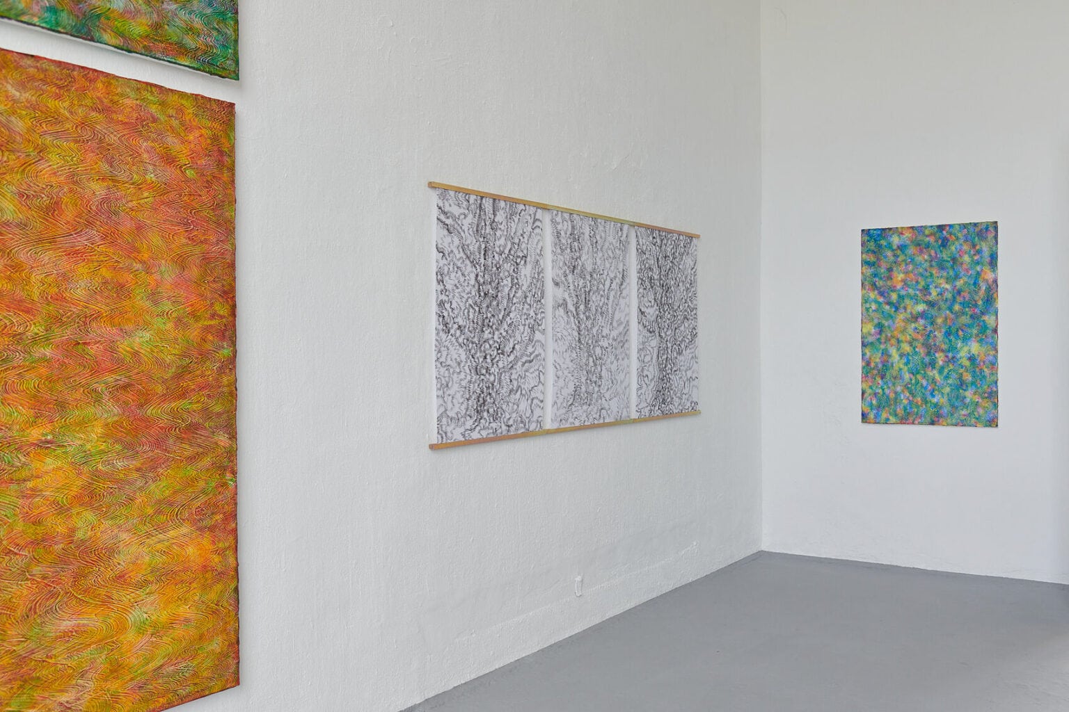 Foto: Ein Raum mit weißen Wänden und grauem Boden. An der Wand hängen Leinwand und Papierarbeiten im Mittel- und Großformat.