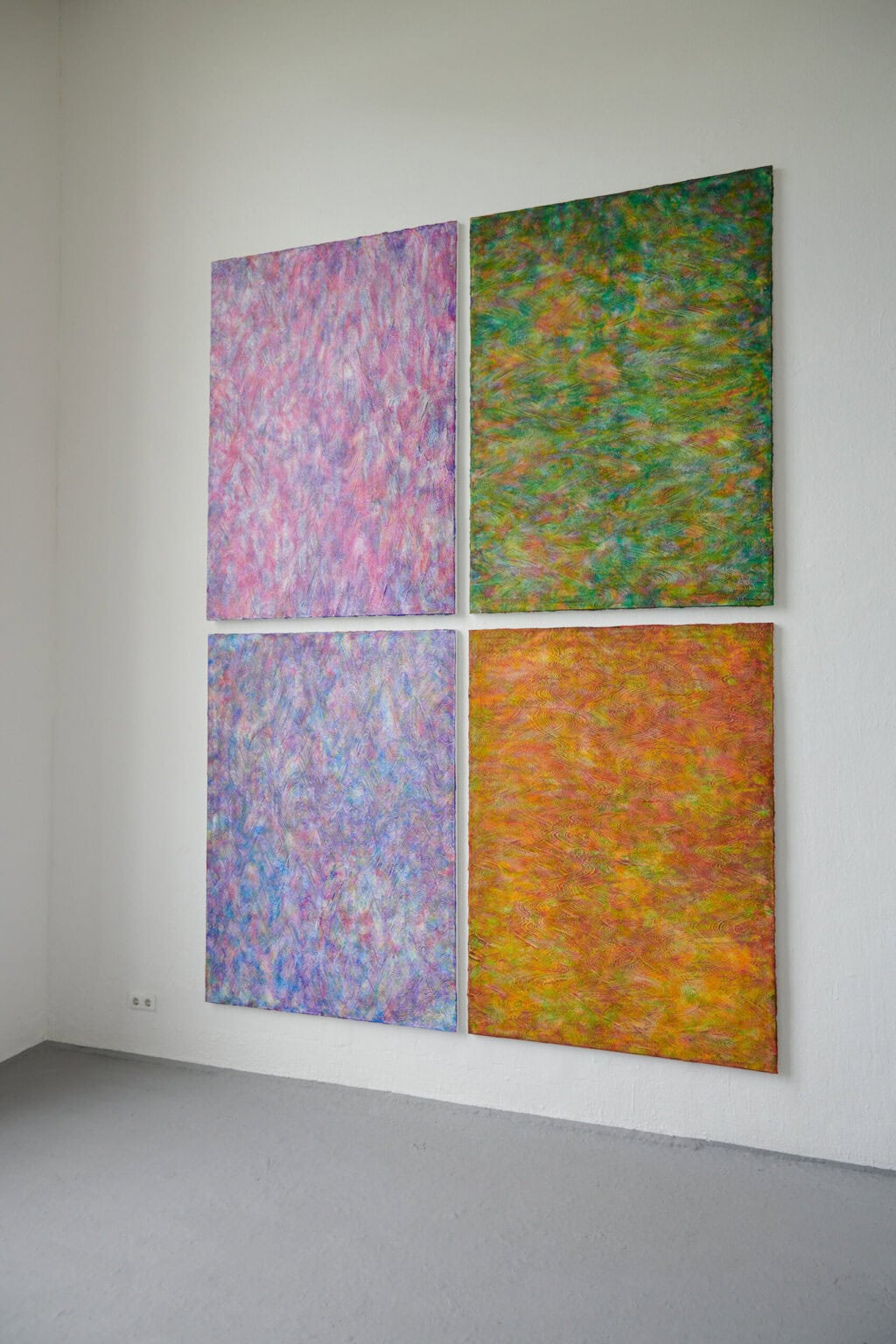 Foto: Ein Raum mit weißen Wänden und grauem Boden. An einer der Wände hängt eine Gruppe aus vier Leinwandarbeiten im gleichen Format.