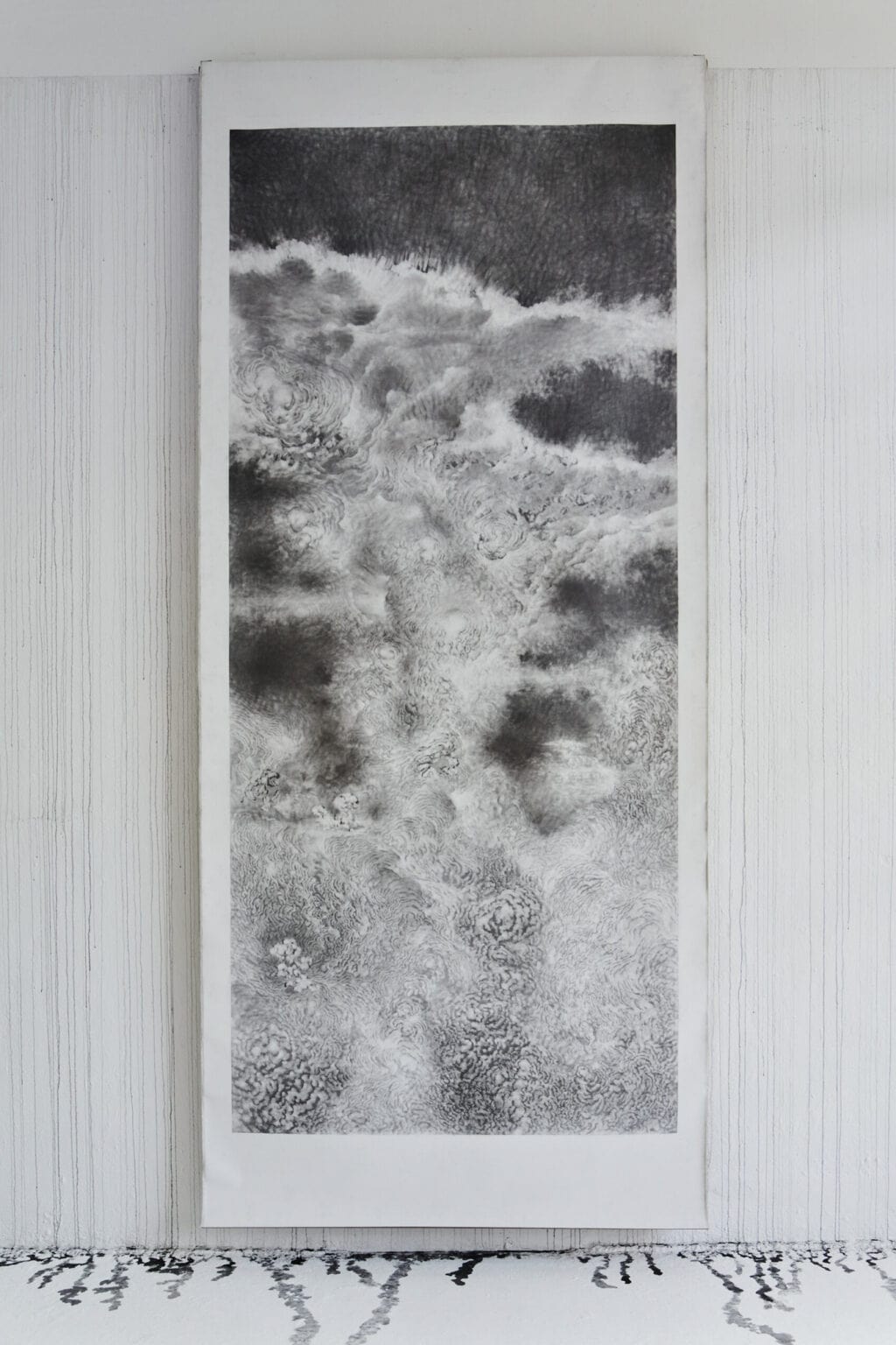 Foto: Ein Raum mit weißen Wänden und grauem Boden. Über die gesamte Länge der weißen Wand ziehen sich Laufspuren schwarzer Farbe, die beim Auftreffen auf dem Boden kleine Lachen bilden. Der Streifen des Bodens, auf dem sich die Lachen abzeichnen, ist im unterschied zum Rest des Bodens in einem Wandnahen Streifen ebenfalls weiß gestrichen. An der Wand hängt eine großformatige, monochrome Arbeit auf Papier.