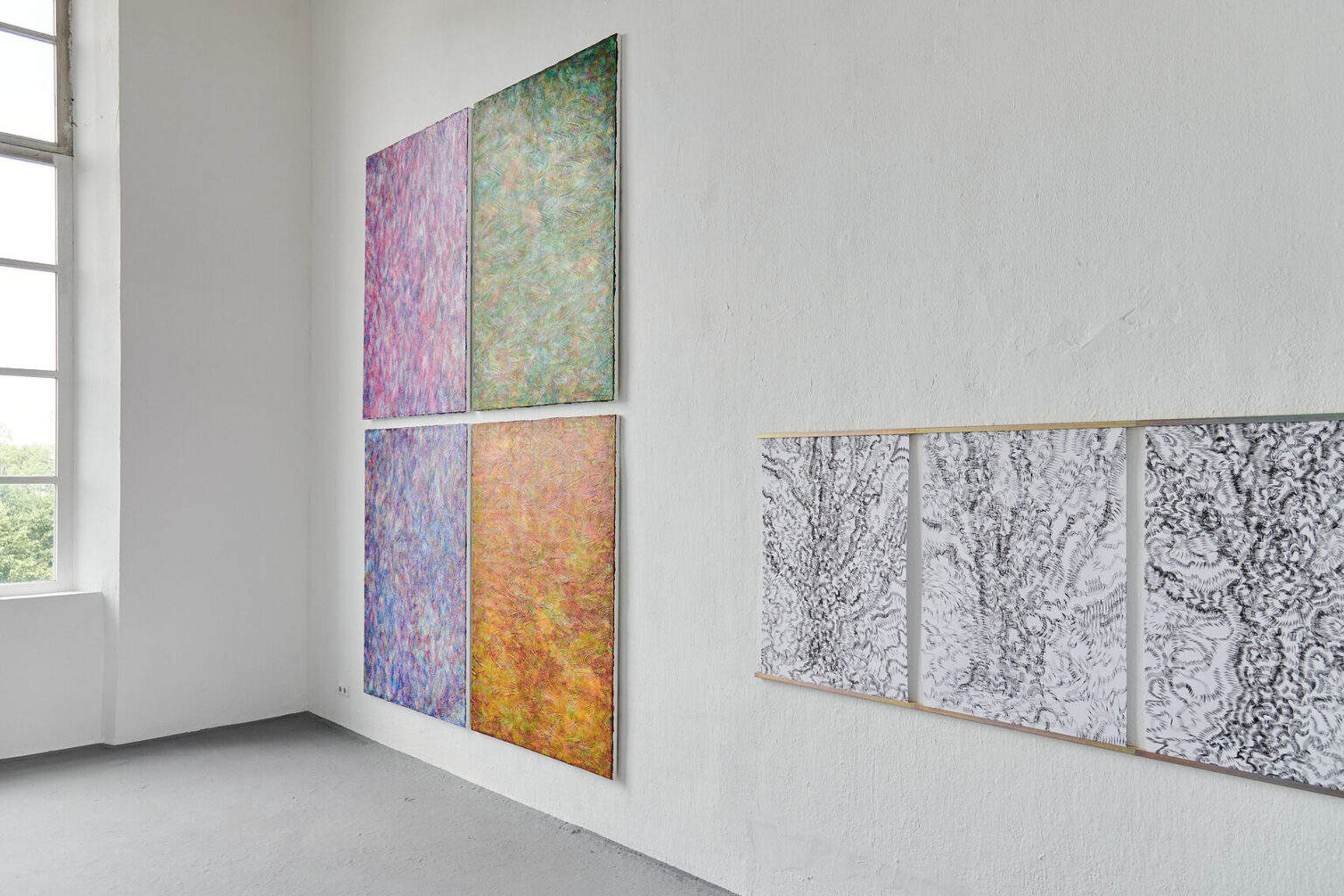 Foto: Ein Raum mit weißen Wänden und grauem Boden. An einer der Wände hängt eine Gruppe aus vier Leinwandarbeiten im gleichen Format. Daneben hängen drei Papierarbeiten, die durch eine Leiste miteinander verbunden und an der Wand fixiert sind.