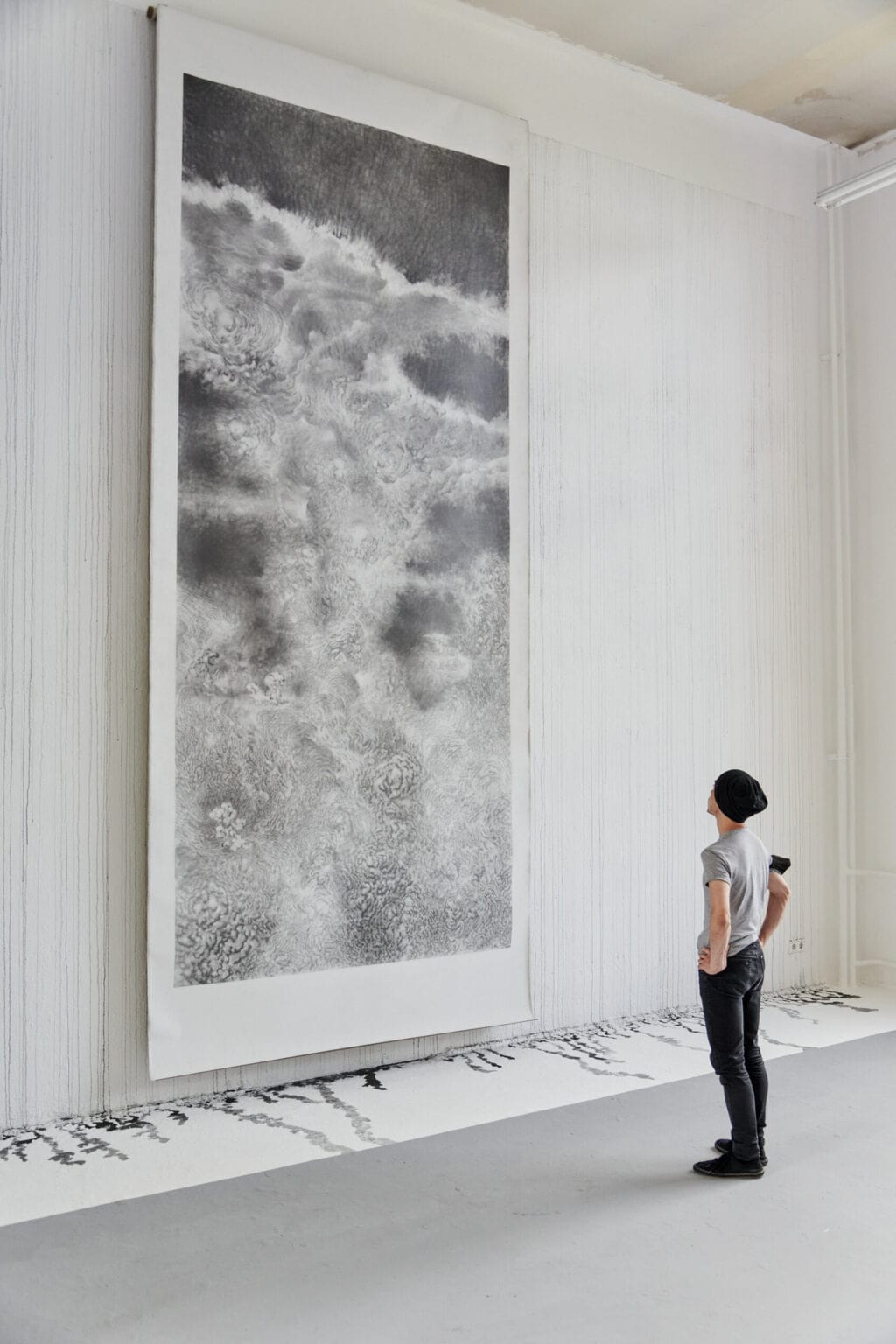 Foto: Ein Raum mit weißen Wänden und grauem Boden. Über die gesamte Länge der weißen Wand ziehen sich Laufspuren schwarzer Farbe, die beim Auftreffen auf dem Boden kleine Lachen bilden. Der Streifen des Bodens, auf dem sich die Lachen abzeichnen, ist im unterschied zum Rest des Bodens in einem Wandnahen Streifen ebenfalls weiß gestrichen. An der Wand hängt eine großformatige, monochrome Arbeit auf Papier. Im Raum steht ein Mensch.