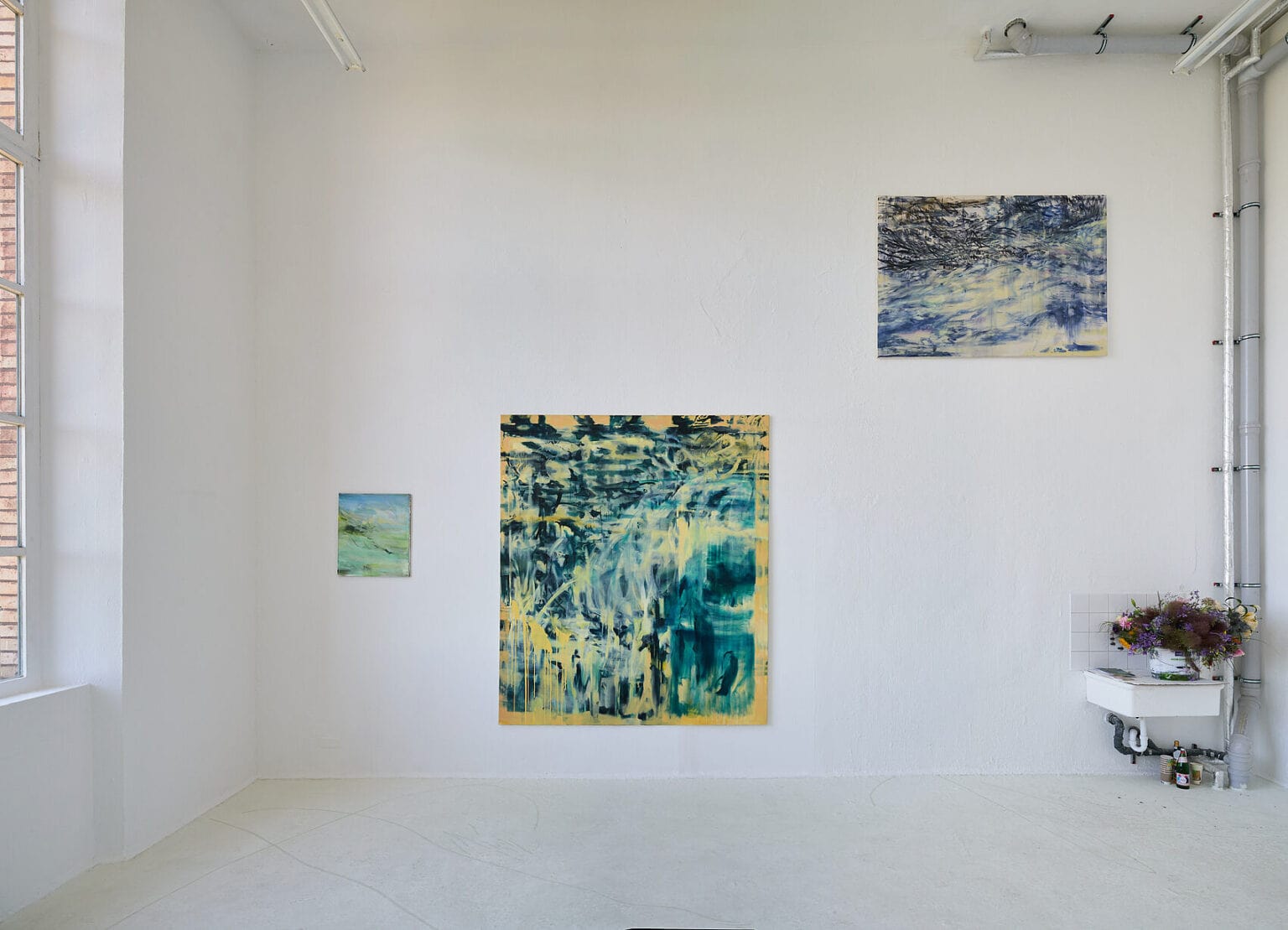 Foto: Ansicht eines Raumes mit weißen Wänden, hellgrauem Boden und einem Fenster. An der Wand befindet sich ein Waschbecken, in dem sich Blumen befinden. An der Wand hängen, auf unterschiedlichen Höhen, Leinwandarbeiten im Klein-, Mittel- und Großformat.