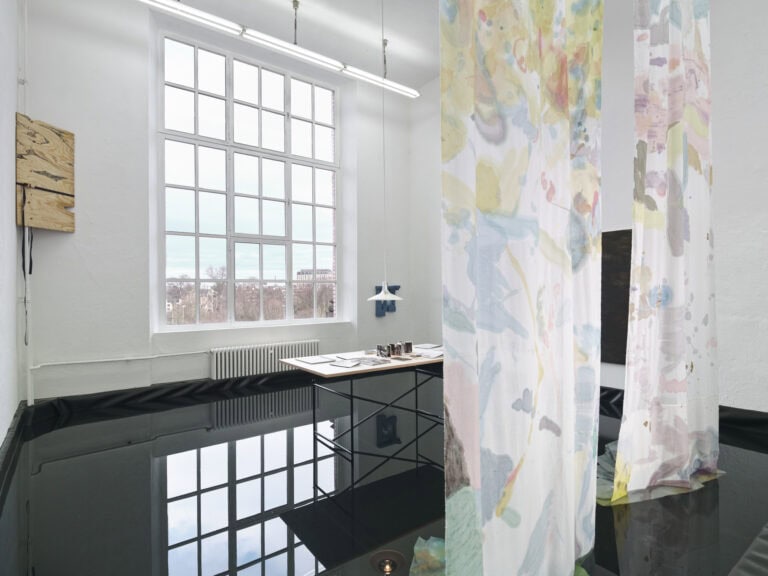 Foto: Ansicht eines Raumes mit weißen Wänden und einem großen Fenster. Der Boden und ein Teil der Wand sind mit schwarzer Folie ausgekleidet. Innerhalb der Folie befindet sich einige Zentimeter tiefes Wasser. Die Oberfläche das Wassers spiegelt den Raum. Ein Tisch steht im Wasser. Auf dem Tisch liegen verschiedene Objekte und eine Lampe schwebt über ihm. Von der Decke hängen farbig gemusterte Textilien herab. An den Wänden befinden sich Objekte.