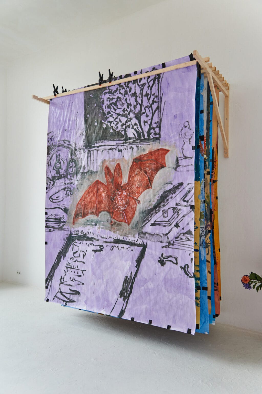 Foto: Ein Raum mit weißen Wänden und weißem Boden. Oben an der Wand befindet sich eine Holzkonstruktion, an der große Papierarbeiten hintereinander aufgehangen sind. Bloß die vorderste Arbeit ist gut zu sehen.