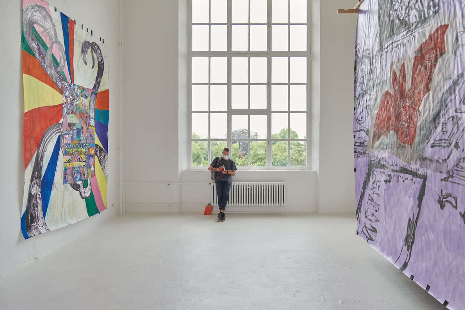 Foto: Ein Raum mit weißen Wänden, hellgrauem Boden und einem großen Fenster, vor dem ein Mensch lehnt. An der linken Wand des Raumes hängt eine großformatige Papierarbeit, die mit schwarzem Tape an der Wand befestigt ist. Rechts ist eine weitere große Papierarbeit zu sehen, die an einer Holzkonstruktion hängt.