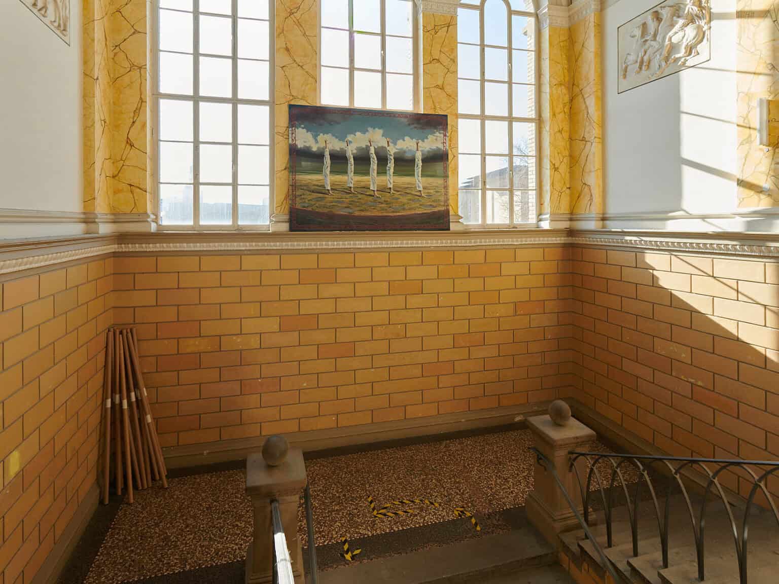 Foto: Ansicht eines Treppenaufgangs mit hohen Wänden und großen Bogenfenstern. Über einem der Fenster hängt eine großformatige Malerei. Licht Fällt durch die Fenster in das Gebäude.