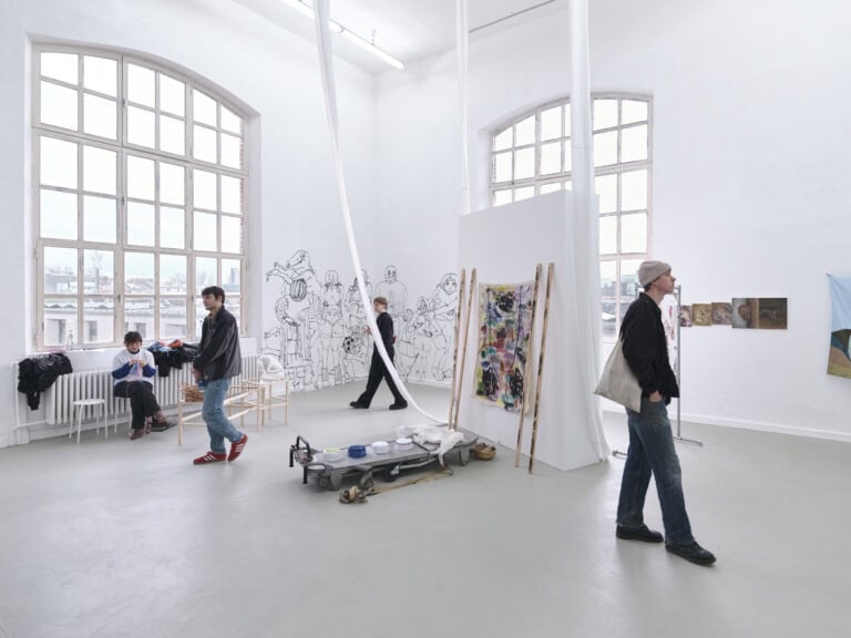 Foto: Ansicht eines Raumes mit weißen Wänden, grauem Boden und zwei Bogenfenstern. An den Wänden befinden sich Objekte und direkt auf die Wand aufgebrachte Malereien. Im Raum stehen Objekte. Menschen bewegen sich durch den Raum und sitzen auf Stühlen. Eine Stellwand ist im Raum aufgestellt. Stoffe hängen von der Decke auf den Boden.