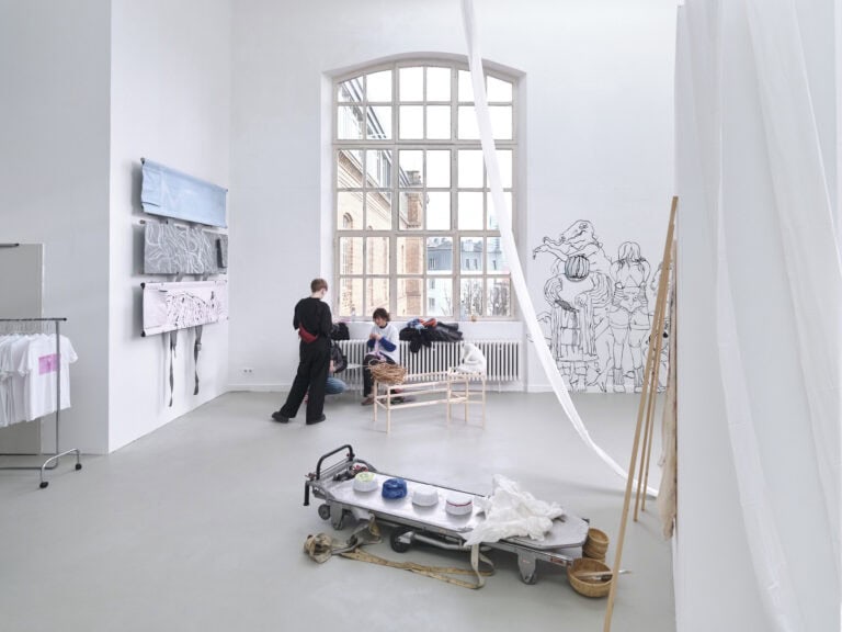 Foto: Ansicht eines Raumes mit weißen Wänden, grauem Boden und einem Bogenfenster. An den Wänden befinden sich Objekte und direkt auf die Wand aufgebrachte Malereien. Im Raum stehen Objekte. Vor dem Fenster stehen und sitzen Menschen.