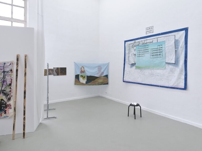 Foto: Aufnahme eines Raumes mit weißen Wänden und grauem Boden. An den Wänden befinden sich flache Objekte. Im Raum steht eine Stellwand, an der Objekte hängen und lehnen.