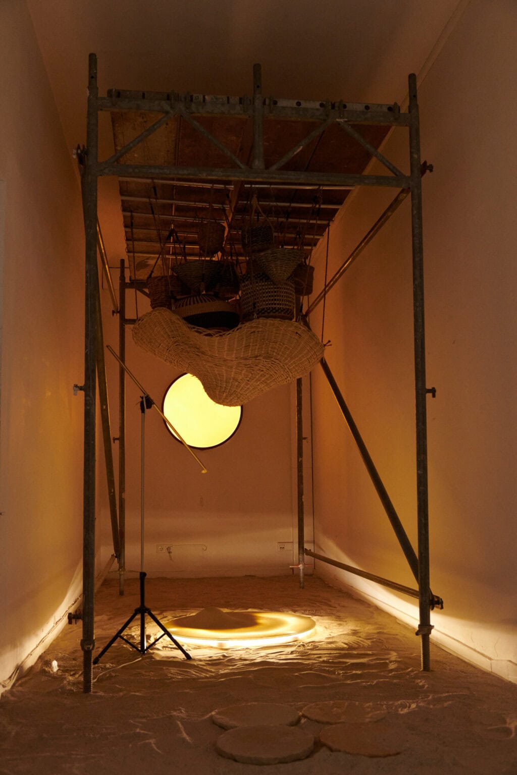 Foto: Ansicht eines schmalen Raumes. Der Boden das Raumes ist mit einer sandigen Masse bedeckt, auf der runde, flache Objekte liegen, auf denen wiederum Sand liegt. Eines der runden Objekte leuchtet und wirft ein warmes, gelbliches Licht in den Raum. Im Raum ist ein Metallgerüst aufgebaut, dass wenig schmaler und höher ist als der Raum selbst. Oben auf dem Gerüst liegen Holzplatten, von denen verschiedengroße geflochtene Objekte hängen. Unter dem Gerüst steht ein Ständer. In die hintere Wand des Raumes ist ein rundes, gelbes Objekt eingelassen.