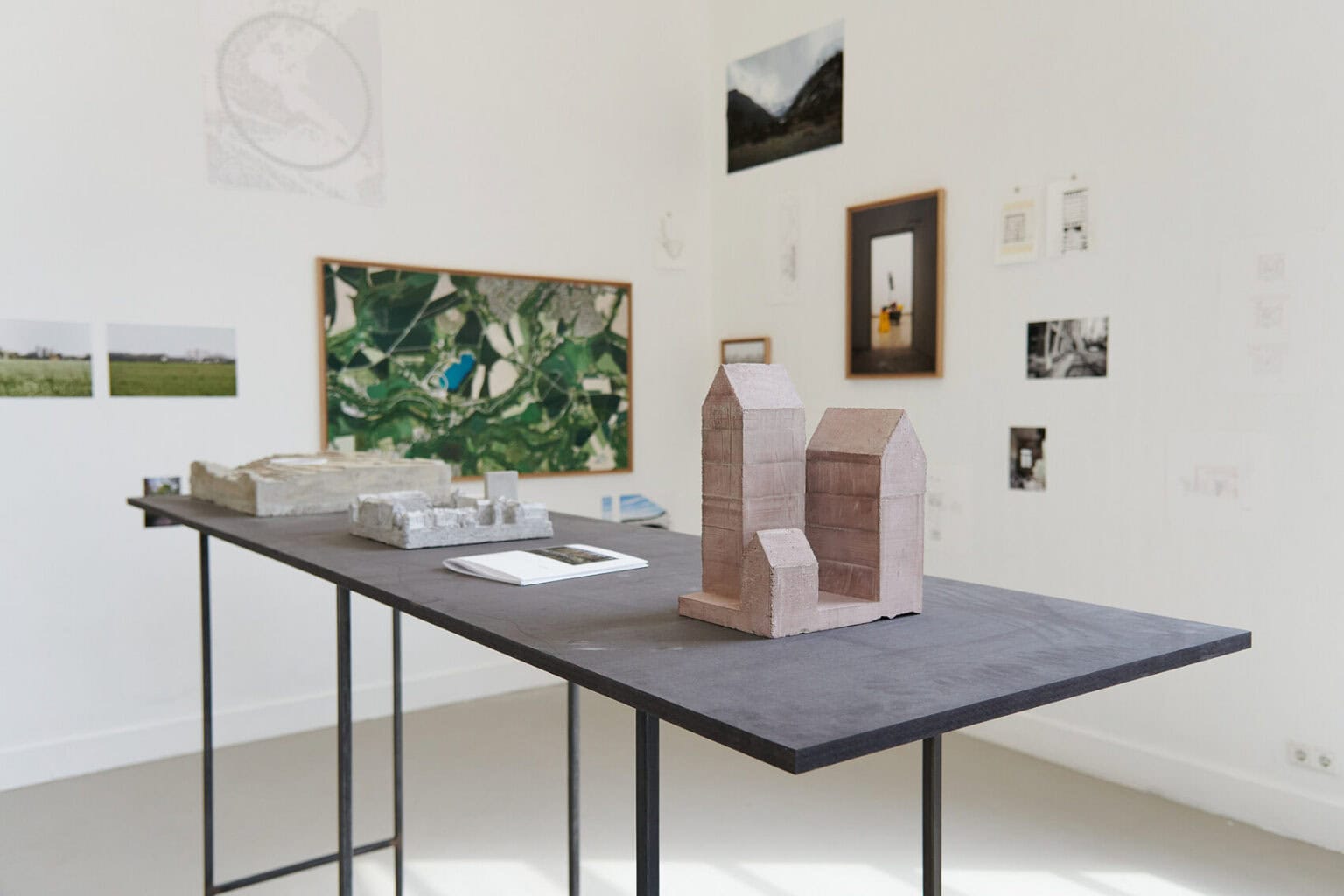 Foto: Architektonische Modelle aus unterschiedlichen Materialien auf einem schwarzen Tisch. An den Wänden befinden sich gerahmt und ungerahmte, flache Objekte und Klein- und Mittelformaten.