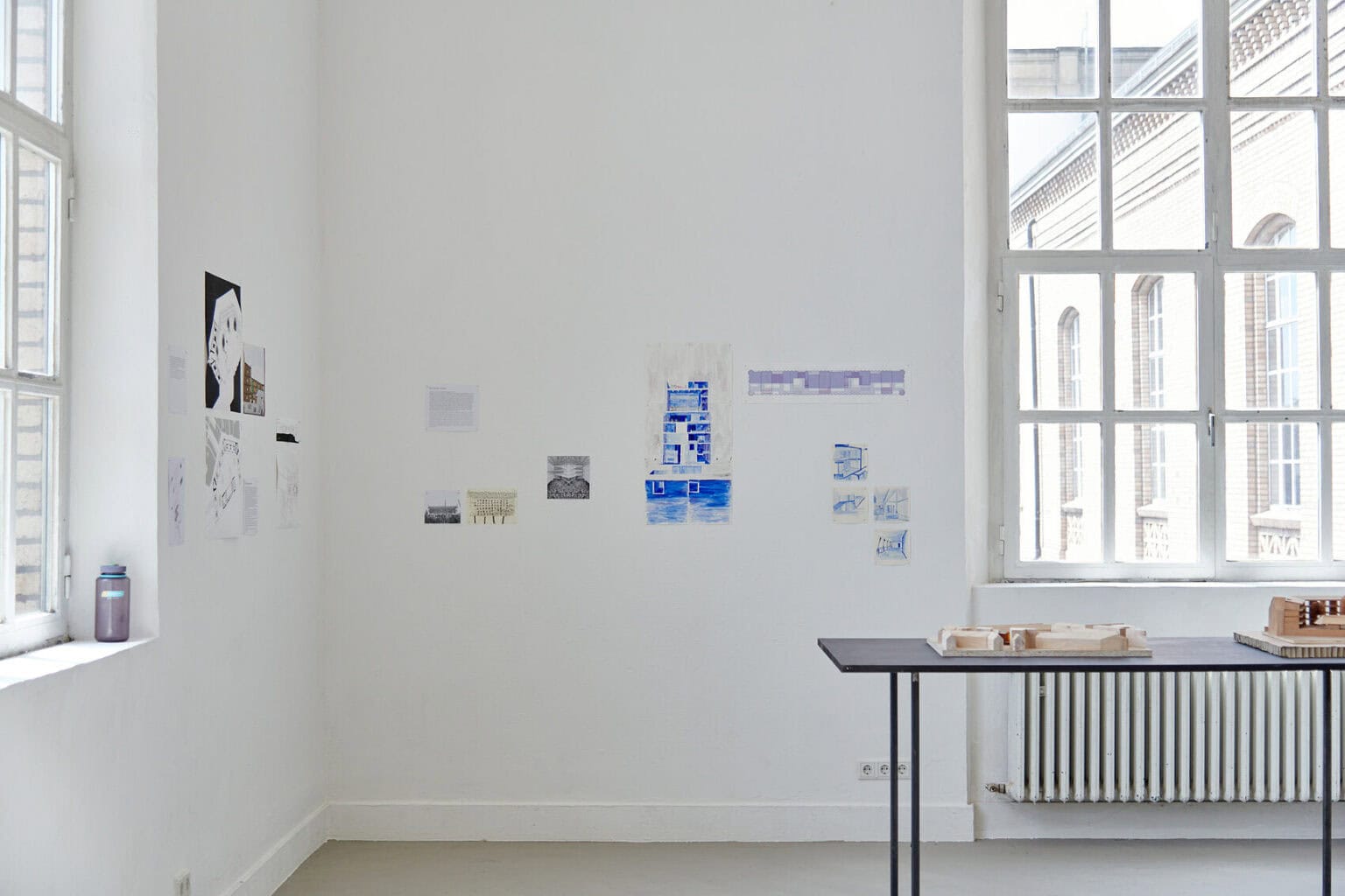 Foto: Ein Raum mit weißen Wänden, grauem Boden und zwei Fenstern. An den Wänden befinden sich kleinformatige Papierarbeiten. Im Raum steht ein Tisch, auf dem sich Objekte befinden.