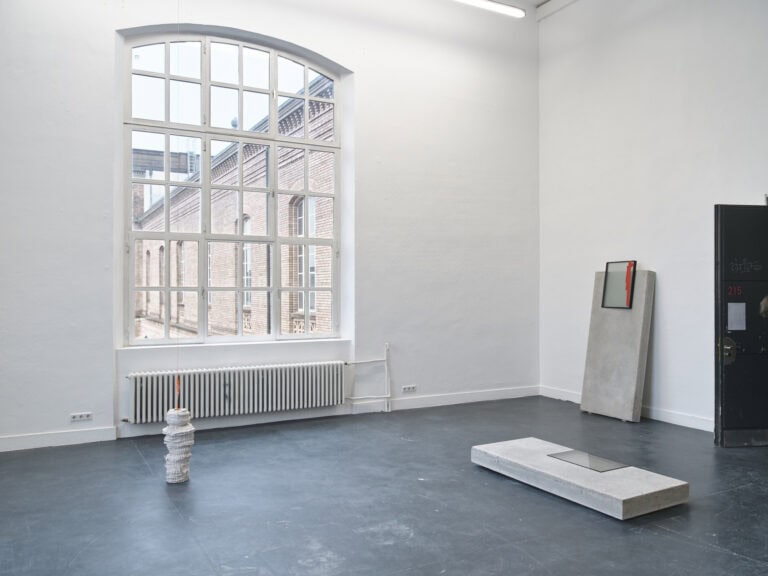 Foto: Aufnahme eines Raums mit weißen Wänden, dunklem Boden und einem Bogenfenster. Im Raum befinden sich stehend und liegend Skulpturen. Weitere von Ihnen sind an die Wände gelehnt.