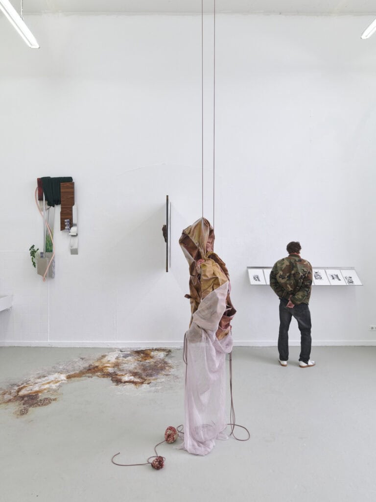 Foto: Ansicht eines Raumes mit weißen Wänden und grauen Boden. Der Boden ist stellenweise mit einer Farbfläche versehen. An den Wänden hängen Objekte. Im Raum schwebt eine Skulptur, die mit Fäden versehen ist, die aus dem Bildfeld reichen. Ein Mensch betrachtet eines der Objekte in Nahsicht.