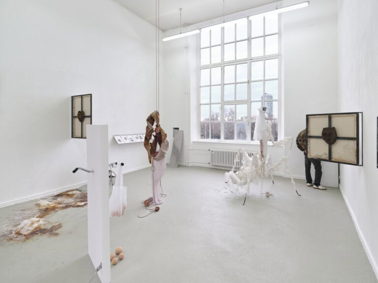 Foto: Ansicht eines Raumes mit weißen Wänden, grauem Boden und einem großen Fenster. An den Wänden und im Raum befinden sich Objekte, die auf dem Boden stehen, von der Decke hängen oder an der Wand montiert sind. Der Boden ist stellenweise mit einer Farbfläche versehen. Hinter einem der Objekte steht ein Mensch.