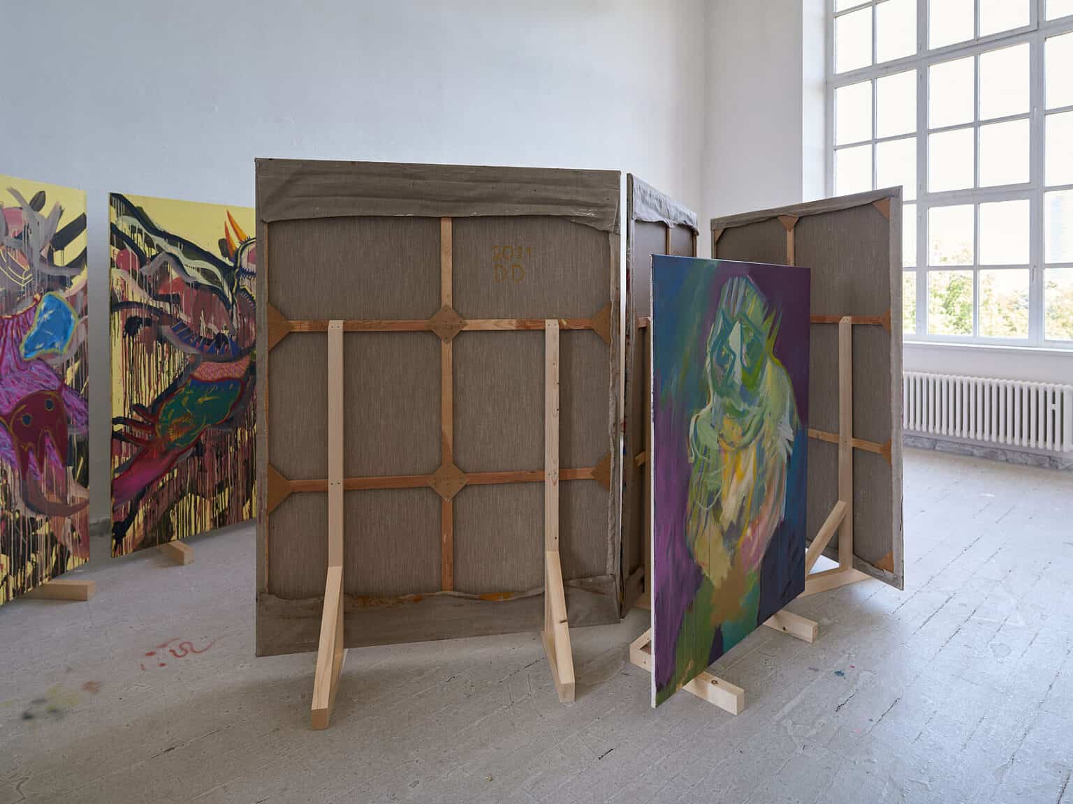 Foto: Ansicht eines Raumes mit weißen Wänden, grauem Dielenboden und einem großen Fenster. Im Raum stehen mehrere groß- und mittelformatige Leinwände auf Holzkonstruktionen.