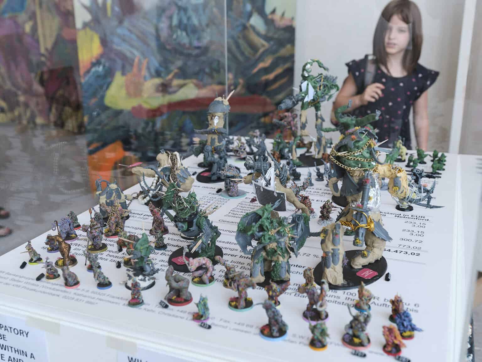 Foto: Ansicht von einer Vielzahl kleiner Objekte auf mit Schrift versehenem Untergrund in einer Vitrine. Im Hintergrund ist ein Mensch zu erkennen.