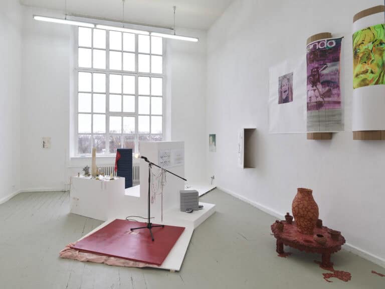 Foto: Ansicht eines Raumes mit weißen Wänden, grünlich-grauem Dielenboden und einem großen Fenster. An den Wänden und im Raum befinden sich Objekte, manche von ihnen stehen auf einem verschachtelte, weißen Podest.