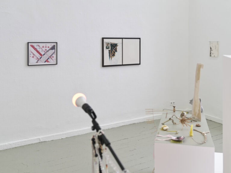 Foto: Ansicht eines Raumes mit weißen Wänden und grau-grünem Dielenboden. Vorne im Bild eine unscharfe Skulptur. Dahinter eine längliche Plattform mit Objekten. An der Wand hängen Arbeiten in Klein- und Mittelformaten.