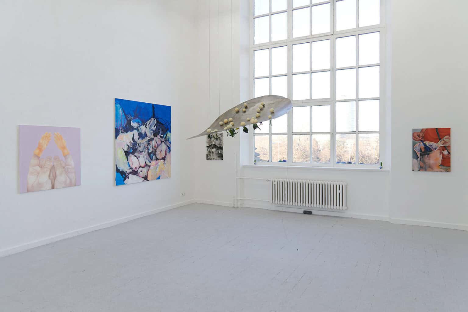 Foto:Ansicht eines Raumes mit weißen Wänden, grauem Dielenboden und einem großen Fenster. An den Wänden hangen Malereien in Klein- und Mittelformaten. Von der Decke hängt, an Seilen befestigt, ein flaches Objekt aus unterschiedlichen Materialien.