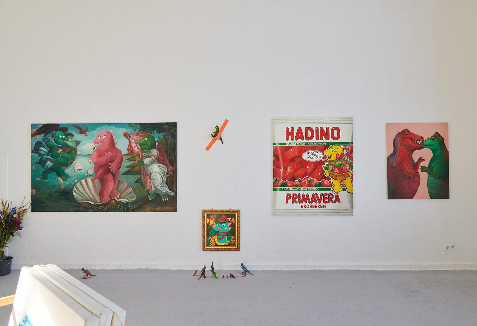 Foto: Ansicht eines Raume mit weißen Wänden und grauem Boden. An der Wand befinden sich, auf unterschiedlicher Höhe, Malereien. Zwischen den Arbeiten befindet sich ein an die Wand getapetes Objekt. Vor der Wand stehen mehrere Objekt und ein Blumenstrauß in einer Vase auf dem Boden.