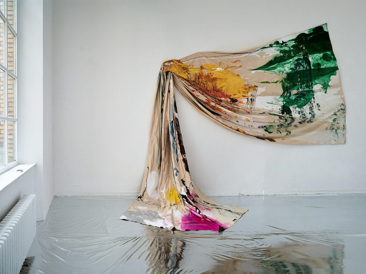 Foto: Ansicht eines Raumes mit weißen Wänden und Fenster. Der Boden ist mit einem silbernen, reflektierendem Material ausgelegt. An der Wand ist ein mit Farbe bearbeitetes Textil horizontal montiert, dass Falten wirft und auf das Material am Boden des Raumes hängt.