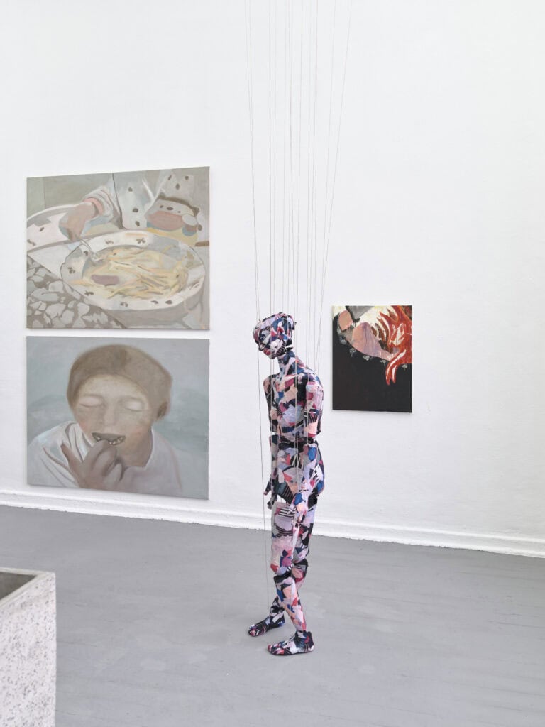 Foto: Ansicht eines Raumes mit weißen Wänden und grauem Boden. An den Wänden befinden sich groß- und mittelformatige Malereien. Im Raum befindet sich eine Skulptur. An die Skulptur sind Fäden angebracht, die aus dem Bildraum hinaus laufen.