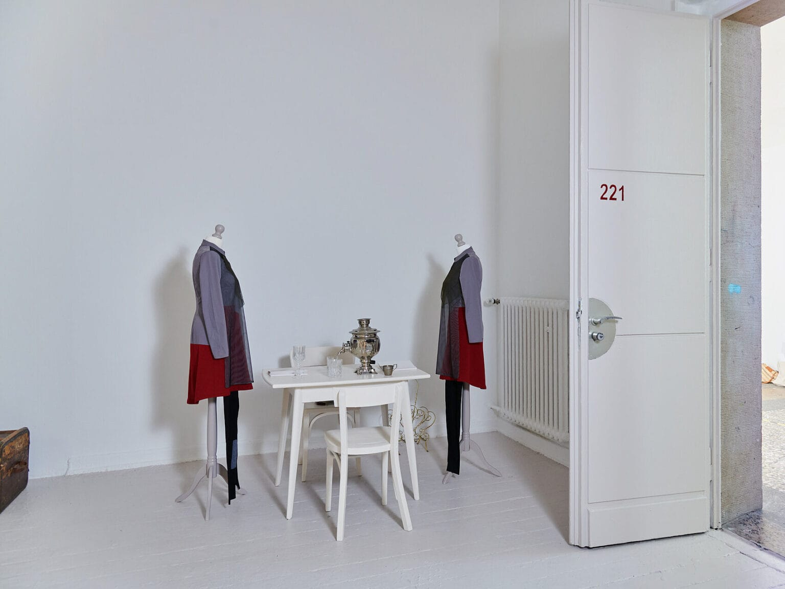 Foto: Ansicht eines Raumes mit weißen Wänden und beigem Boden. Im Raum steht ein weißer Tisch, auf dem sich Hefte, Gläser und ein Samowar befinden. Am Tisch stehen zwei weiße Stühle und zwei bekleidete Schneiderpuppen. Auf dem Boden befinden sich zwei Objekte.