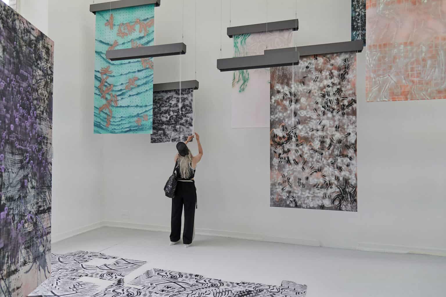 Foto: Ein Raum mit weißen Wänden und grauem Dielenboden. Von der Decke hängen Fäden, an denen flache, herabhängenden Objekte befestigt sind. Auf dem Boden liegt eine Gruppe flacher, gemusterter Objekte. Links im Raum steht eine großformatige Leinwandarbeit. Im Raum steht eine Person, die mit einer der Arbeiten interagiert.