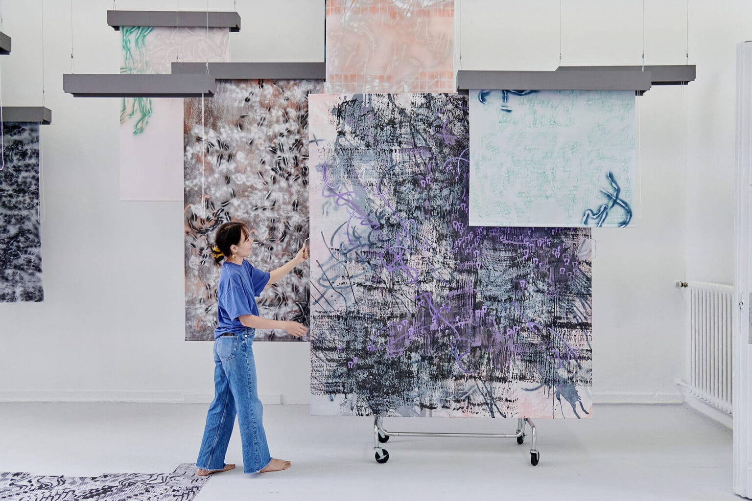 Foto: Ein Raum mit weißen Wänden und grauem Dielenboden. Von der Decke hängen Fäden, an denen flache, herabhängenden Objekte befestigt sind. Auf dem Boden liegt eine Gruppe flacher, gemusterter Objekte. Eine Person schiebt eine großformatige Leinwand durch den Raum, die auf einer Konstruktion aus Rollen befestigt ist.