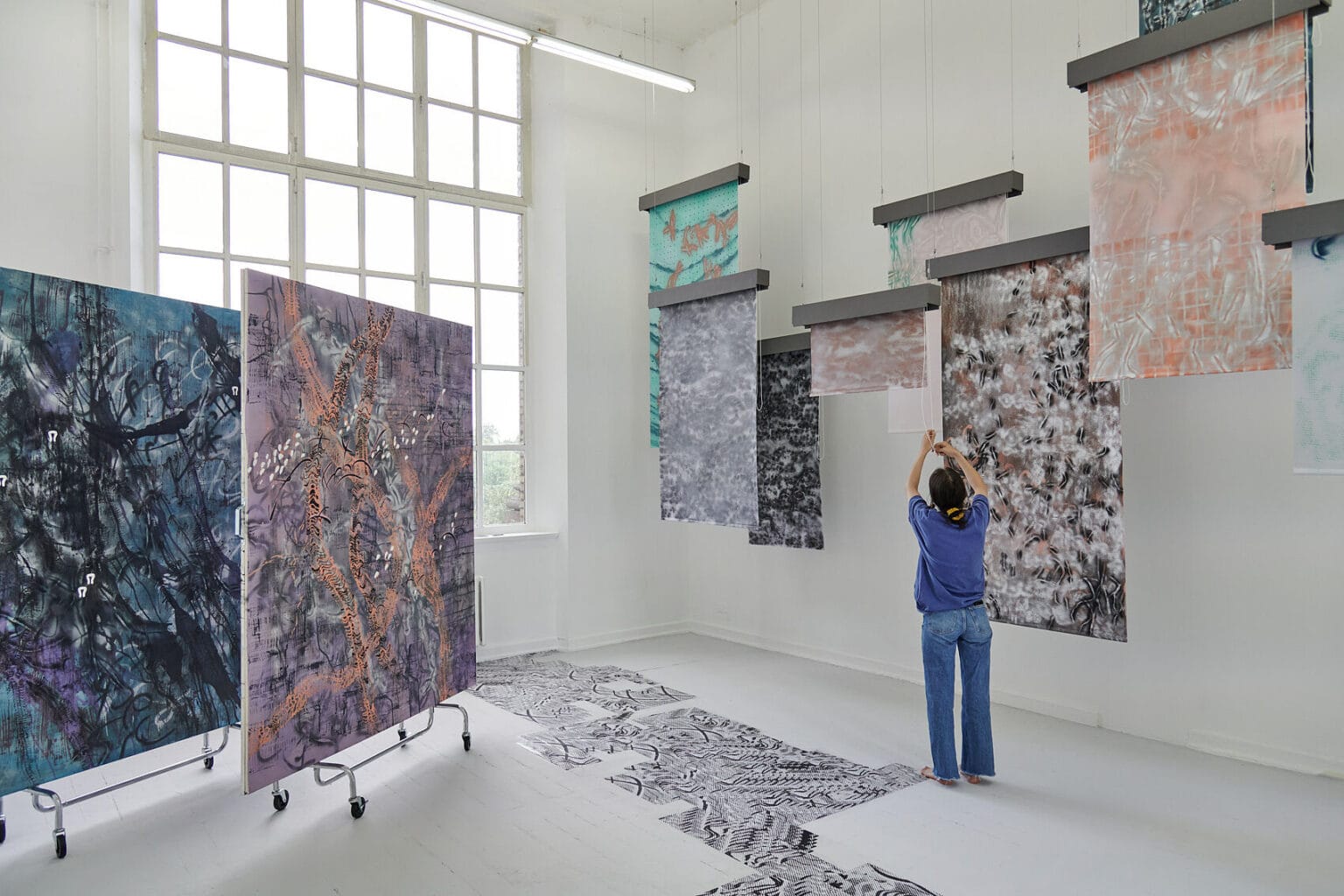 Foto: Ein Raum mit weißen Wänden, grauem Dielenboden und einem großen Fenster. Von der Decke hängen Fäden, an denen flache, herabhängenden Objekte befestigt sind. Auf dem Boden liegt eine Gruppe flacher, gemusterter Objekte. Links im Raum stehen zwei großformatige Leinwandarbeiten, die auf einem mit Rollen versehenen Gerüst stehen. Im Raum steht eine Person, die mit einer der Arbeiten interagiert.