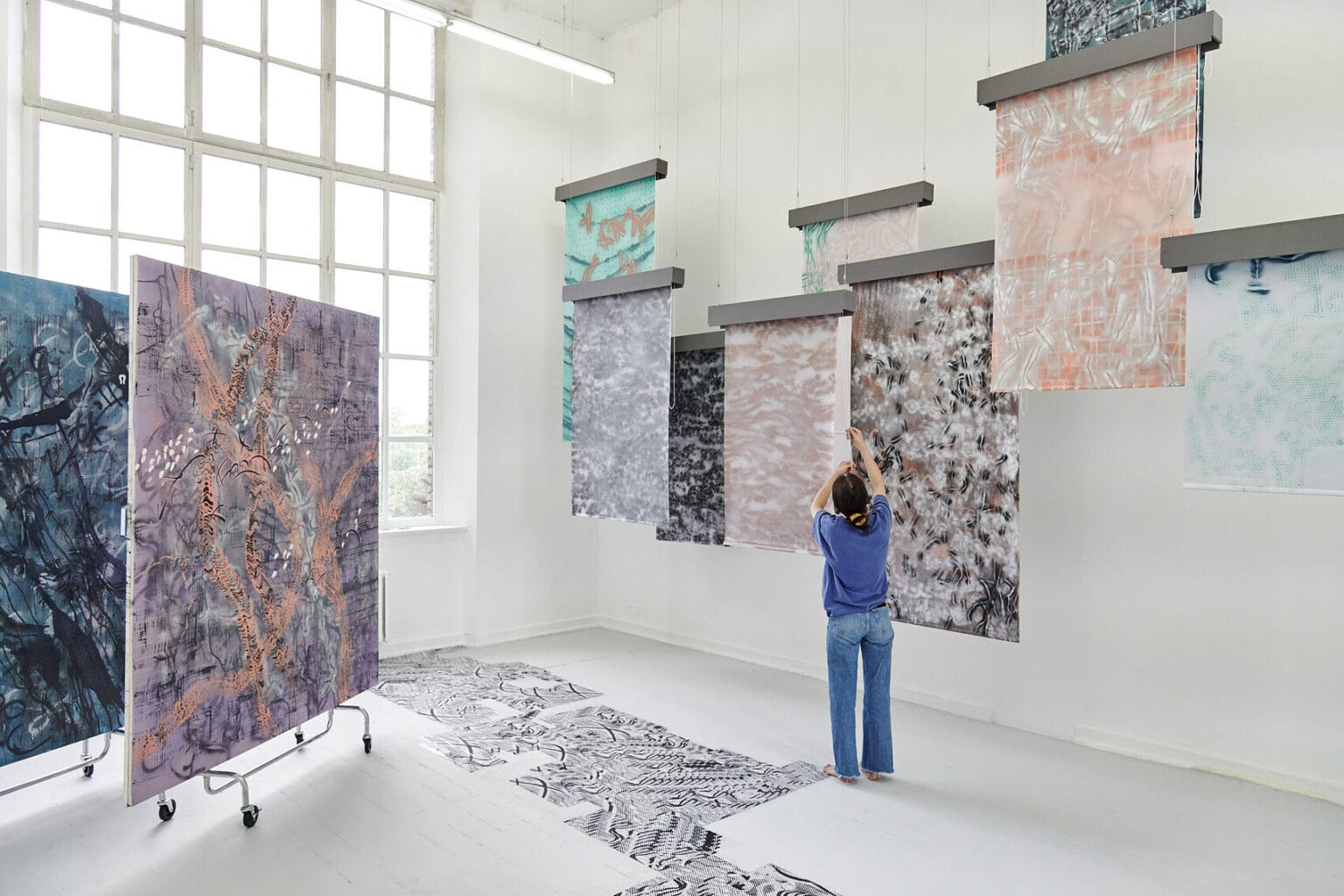 Foto: Ein Raum mit weißen Wänden, grauem Dielenboden und einem großen Fenster. Von der Decke hängen Fäden, an denen flache, herabhängenden Objekte befestigt sind. Auf dem Boden liegt eine Gruppe flacher, gemusterter Objekte. Links im Raum stehen zwei großformatige Leinwandarbeiten, die auf einem mit Rollen versehenen Gerüst stehen.Im Raum steht eine Person, die mit einer der Arbeiten interagiert.